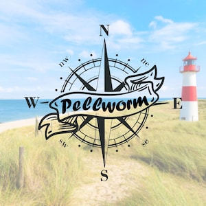 Könnte beinhalten: Ein schwarz-weißer Kompass mit dem Wort "Pellworm" auf einem blauen Band. Der Kompass ist vor dem Hintergrund eines Strandes mit einem Leuchtturm in der Ferne platziert.