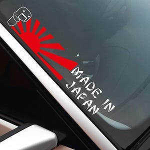 Könnte beinhalten: Ein weißer Vinyl-Aufkleber mit dem Text "Made in Japan" und einer roten aufgehenden Sonne Grafik mit einem Cartoon-Monster auf der linken Seite.