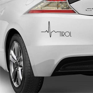 Könnte beinhalten: Ein weißes Auto mit einem schwarzen Aufkleber auf der hinteren Stoßstange. Der Aufkleber ist ein stilisiertes Herzschlag mit dem Wort "TIROL" darunter.
