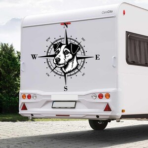 Può includere: Un camper bianco con un design a bussola in bianco e nero con la faccia di un cane. La bussola punta verso i punti cardinali: Nord, Est, Sud e Ovest. Il camper ha luci rosse e arancioni.