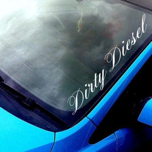 Op de afbeelding: Een blauwe auto met een witte vinyl sticker op de voorruit met de tekst "Dirty Diesel".