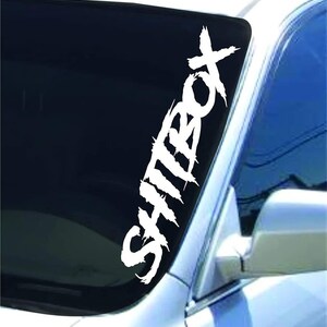 Könnte beinhalten: Ein weißer Vinyl-Aufkleber mit dem Wort "Shitbox" in einer stilisierten Schrift. Der Aufkleber ist auf die Windschutzscheibe eines Autos geklebt.