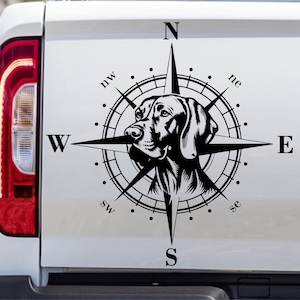 Op de afbeelding: Zwart-wit afbeelding van een hondenhoofd in een kompasroosontwerp. Het kompas wijst naar het noorden, zuiden, oosten en westen, met extra richtingaanwijzers. De afbeelding is waarschijnlijk een sticker.