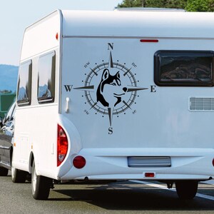 Könnte beinhalten: Ein weißer Wohnwagen mit einem schwarzen Kompass-Aufkleber, der den Kopf eines Huskys zeigt. Der Kompass zeigt nach Norden, Süden, Osten und Westen.