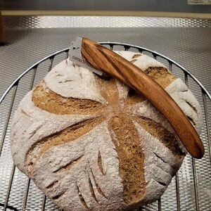 May include: A loaf of sourdough bread with a decorative scoring pattern, resting on a wire rack. A wooden handled razor blade is resting on top of the bread.