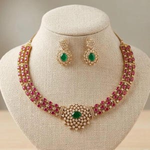 May include: A gold-toned necklace and earring set featuring three rows of oval-cut red stones. The necklace includes a central circular pendant with a prominent oval green stone surrounded by small clear crystals. Matching earrings feature a single oval green stone.