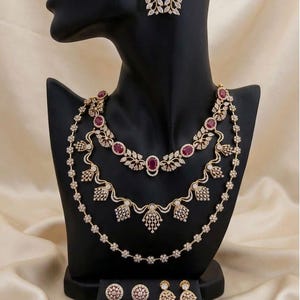 May include: A multi-layered gold-tone necklace featuring oval red stones and clear crystals, paired with matching drop earrings and two additional pairs of small stud earrings, all displayed on a black mannequin bust.