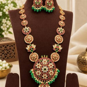May include: A gold-toned necklace and matching earring set featuring intricate circular and lotus-shaped motifs. The jewelry is adorned with red and green stones, clear crystals, and small green beads, creating a vibrant, detailed, and ornate appearance.