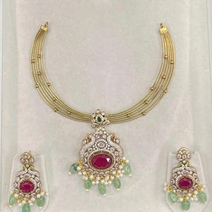 May include: A gold-toned multi-strand necklace featuring a central pendant with a large oval red stone, surrounded by clear crystals, small white pearls, and light green teardrop beads. Matching earrings feature a similar design.
