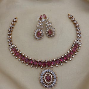 May include: A necklace and matching pair of earrings featuring oval-cut dark pink stones surrounded by small clear white stones. The necklace has a central floral-shaped pendant with a pink stone center and a row of pink stones along the chain.