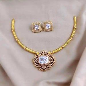 May include: A gold-toned necklace and earring set featuring a flexible snake chain necklace with a large, rectangular, clear-cut center stone surrounded by smaller pavé crystals. The matching stud earrings feature a rectangular clear-cut stone.
