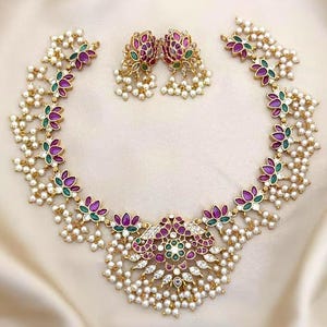 May include: A gold-toned necklace and matching earring set featuring a central peacock motif adorned with pink and green stones, clear crystals, and dangling white pearl-like beads. The necklace chain consists of repeating floral-shaped links.