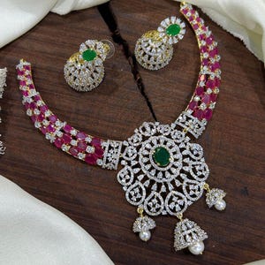 May include: A gold-tone necklace and earring set featuring oval-cut pink stones, clear white crystals, and a central pendant with a green oval stone. The necklace includes three dangling bell-shaped charms with white pearls at the base.