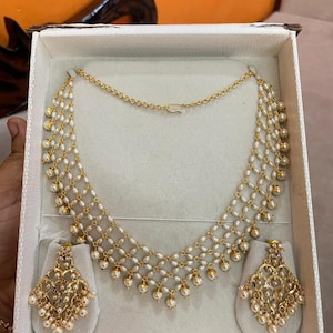 May include: A gold-toned necklace and matching earring set featuring a delicate lattice design crafted from small white pearls. The necklace has a multi-strand mesh structure with dangling pearl accents, and the earrings feature an intricate filigree pattern with pearl embellishments.