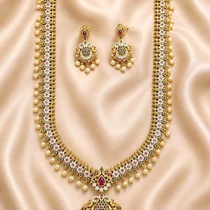 Può includere: Una lunga collana d'oro e un paio di orecchini abbinati caratterizzati da un'intricata lavorazione in metallo, incastonati con piccole pietre bianche, accenti verdi e pietre rosse centrali. Il set è decorato con perle bianche pendenti.