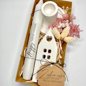 Könnte beinhalten: Eine Geschenkbox gefüllt mit einer weißen Kerze, einem kleinen weißen Haus-Anhänger, einem weißen Kerzenhalter und getrockneten Blumen. Die Kerze trägt den Text "Happy New Home". Das am Haus-Anhänger befestigte Etikett lautet "Zuhause ist, wo die Seele ankommt".
