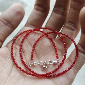 May include: A delicate red beaded necklace with a silver clasp. The necklace is composed of small, round, red beads strung together. The clasp is silver and features a heart-shaped charm. The necklace is displayed on a hand.
