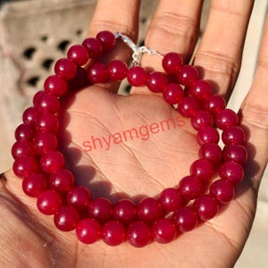 May include: A necklace of deep red, round beads, possibly gemstones, is displayed. The beads are strung together with a silver clasp. The text "shyamgems" is visible on the surface. The necklace is held in an open hand.