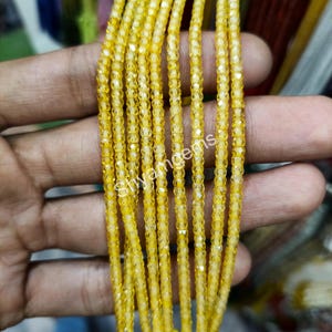 May include: Close-up of strands of yellow gemstone beads. The beads are small, faceted, and strung together, creating a shimmering effect. The text "Shyamgems" is visible on the beads. The beads are held in a hand.