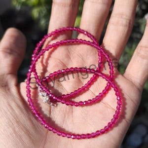 May include: A delicate necklace featuring small, faceted, magenta-colored beads. The necklace is strung together with a silver clasp. The beads are evenly spaced, creating a continuous strand. The necklace is shown on a hand.