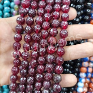 Puede incluir: Primer plano de hileras de cuentas facetadas de piedras preciosas de color rojo intenso. Las cuentas tienen una superficie texturizada y están ensartadas, mostrando un color rico y oscuro. También se ve el texto "shyamgems".