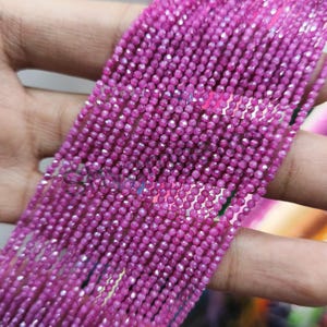 Può includere: Primo piano di più fili di piccole perline sfaccettate color magenta. Le perline sono infilate insieme, creando un aspetto fluido e strutturato. Le perline sono tenute in una mano, mostrando il loro colore e la loro lucentezza. Ideale per la creazione di gioielli.