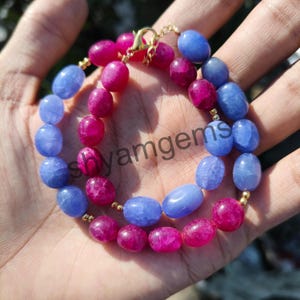May include: A handmade necklace featuring alternating blue and magenta gemstone beads. The necklace is strung on a gold-colored chain and clasp. The beads are oval-shaped and have a polished finish. The necklace is displayed on a hand.