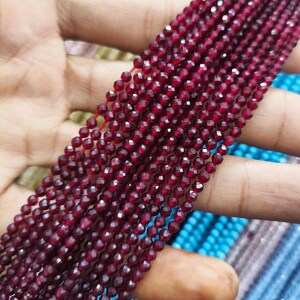 Könnte beinhalten: Nahaufnahme von mehreren Strängen tiefroter Edelsteinperlen. Die kleinen, facettierten Perlen sind aufgefädelt und erzeugen einen schimmernden Effekt. Der Hintergrund zeigt weitere Perlenstränge in Blau- und Türkistönen.