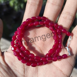 Op de afbeelding: Een ketting bestaande uit kleine, ovale, dieprode kralen. De kralen zijn aan elkaar geregen en vormen een doorlopende lus. Een zilverkleurige sluiting zet de ketting vast. De ketting wordt op een hand getoond. De kralen zijn ongeveer 0,5 cm lang.