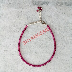 Peut inclure: Un bracelet de perles délicat, de couleur magenta, avec une chaîne et un fermoir argentés. Le bracelet comporte de petites perles rondes et le texte "SHYAMGEMS" en rose. Le bracelet est conçu pour être porté au poignet.