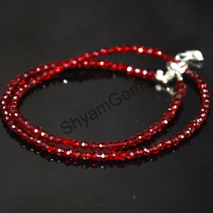 May include: A delicate red gemstone necklace with a silver clasp. The necklace is composed of small, faceted red beads, creating a sparkling effect. The jewelry is displayed on a dark surface.