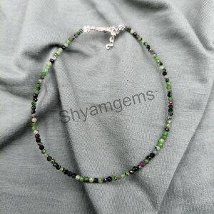 May include: A delicate beaded necklace featuring small, round gemstones in shades of green, black, and maroon. The necklace is strung on a thin, silver-colored chain and is displayed on a light gray fabric. The text "Shyamgems" is visible.