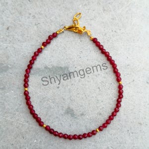 Puede incluir: Una delicada pulsera con pequeñas cuentas facetadas de color rojo intenso, intercaladas con detalles dorados. La pulsera tiene un cierre dorado y el texto "Shyamgems".