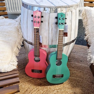 May include: Two ukuleles, one pink and one teal green, are sitting on a white wooden barrel. Both ukuleles have wooden necks and brown strings.
