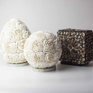 May include: Three decorative spheres made of white seashells, arranged on a white surface. The spheres are in the shape of a flower bud, with each petal made of a single seashell. A small, square box made of brown seashells is behind the spheres.