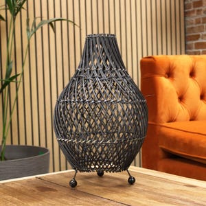 May include: A dark grey woven wicker vase with a bulbous base and a narrow neck. The vase sits on three small, black ball feet. The woven pattern creates a textured surface. The vase is displayed on a wooden table.
