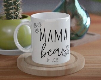 May include: White ceramic mug with black text that reads "Mama Bear Est. 2023" and paw prints around the text.