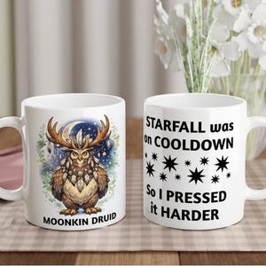 Puede incluir: Dos tazas de cerámica blanca. Una taza presenta un búho con cuernos, una luna creciente y el texto "MOONKIN DRUID". La otra taza tiene el texto "STARFALL was on COOLDOWN, So I PRESSED it HARDER" con gráficos de estrellas.