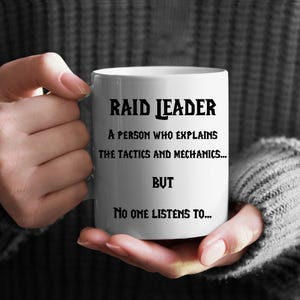 Könnte beinhalten: Eine weiße Keramiktasse mit schwarzem Text, der "RAID LEADER A PERSON WHO EXPLAINS THE TACTICS AND MECHANICS... BUT NO ONE LISTENS TO..." lautet.