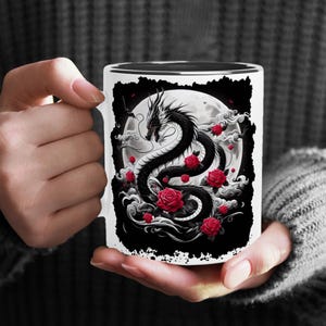 May include: White ceramic mug featuring a black and white illustration of a dragon with red roses surrounding it. The dragon is curled around a full moon.