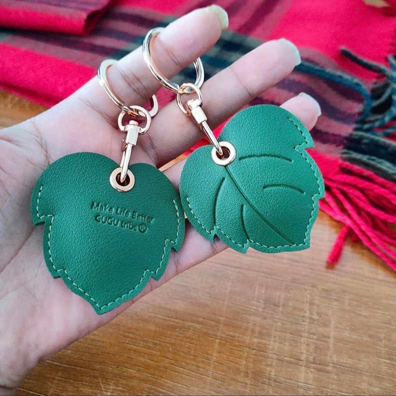 May include: Two green leather leaf-shaped keychains with gold rings and a gold chain. The keychain on the left has the text "Make Life Better CUGU Tribe" on it.
