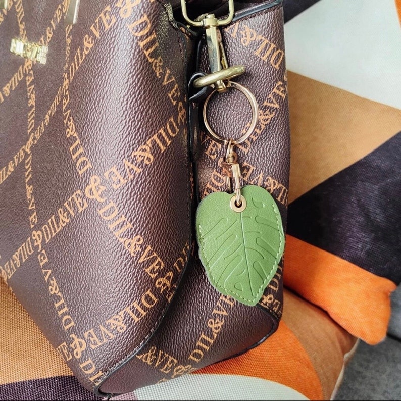 May include: A brown handbag with a gold-colored clasp and a green leaf-shaped keychain. The bag has a repeating pattern of the text "DIL&VE" in a lighter brown color. The keychain is attached to the bag with a gold-colored ring.