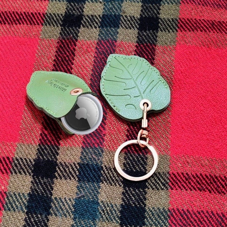 May include: A light green leather leaf-shaped keychain with a gold-colored keyring. The keychain is next to a matching green leather case with a circular tracker inside. The case has text on it.