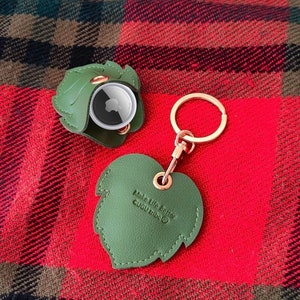 May include: A green leather keychain with a leaf shape and the text "Make life better GUGU tribe" on it. The keychain has a small round compartment with a gold metal lid.
