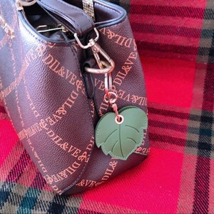 May include: A brown leather handbag with a gold-tone chain strap and a green leaf-shaped keychain. The bag has a repeating pattern of the text "DIL&VE".