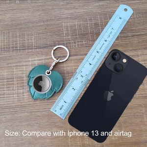 May include: A teal leather keychain shaped like a leaf with a silver ring. The keychain is next to a ruler and an iPhone 13 to show its size.