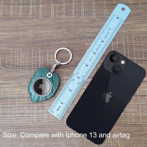 May include: A teal leather AirTag holder in a leaf shape with a silver keyring, a blue ruler, and a black iPhone 13. The ruler shows inches. The text at the bottom says, "Size: Compare with Iphone 13 and airtag."