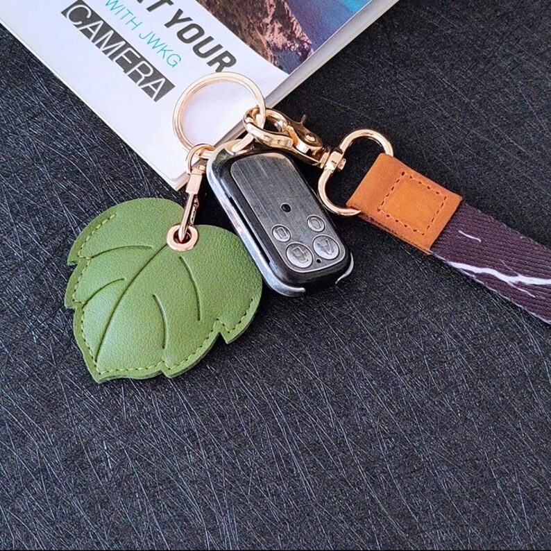 May include: A green leather leaf keychain with a gold ring and a black remote control attached. The keychain is connected to a brown and black striped lanyard with a brown leather patch.
