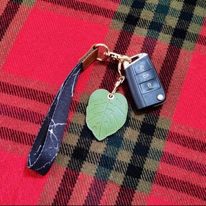 May include: A black and white marble patterned wrist strap keychain with a gold-colored clasp, a green leaf-shaped charm, and a car key. The keychain is on a red and black plaid background.