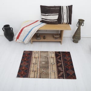 May include: A small, rectangular rug with a beige background and a geometric pattern in shades of red, brown, and black. The rug is on a white floor in front of a wooden bench with two pillows.